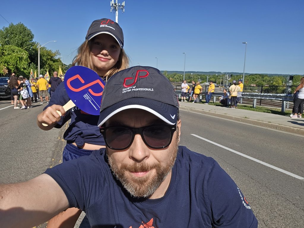 Image of Mike wearing a Society of United Professionals hat. A child is behind him with a Society sign.